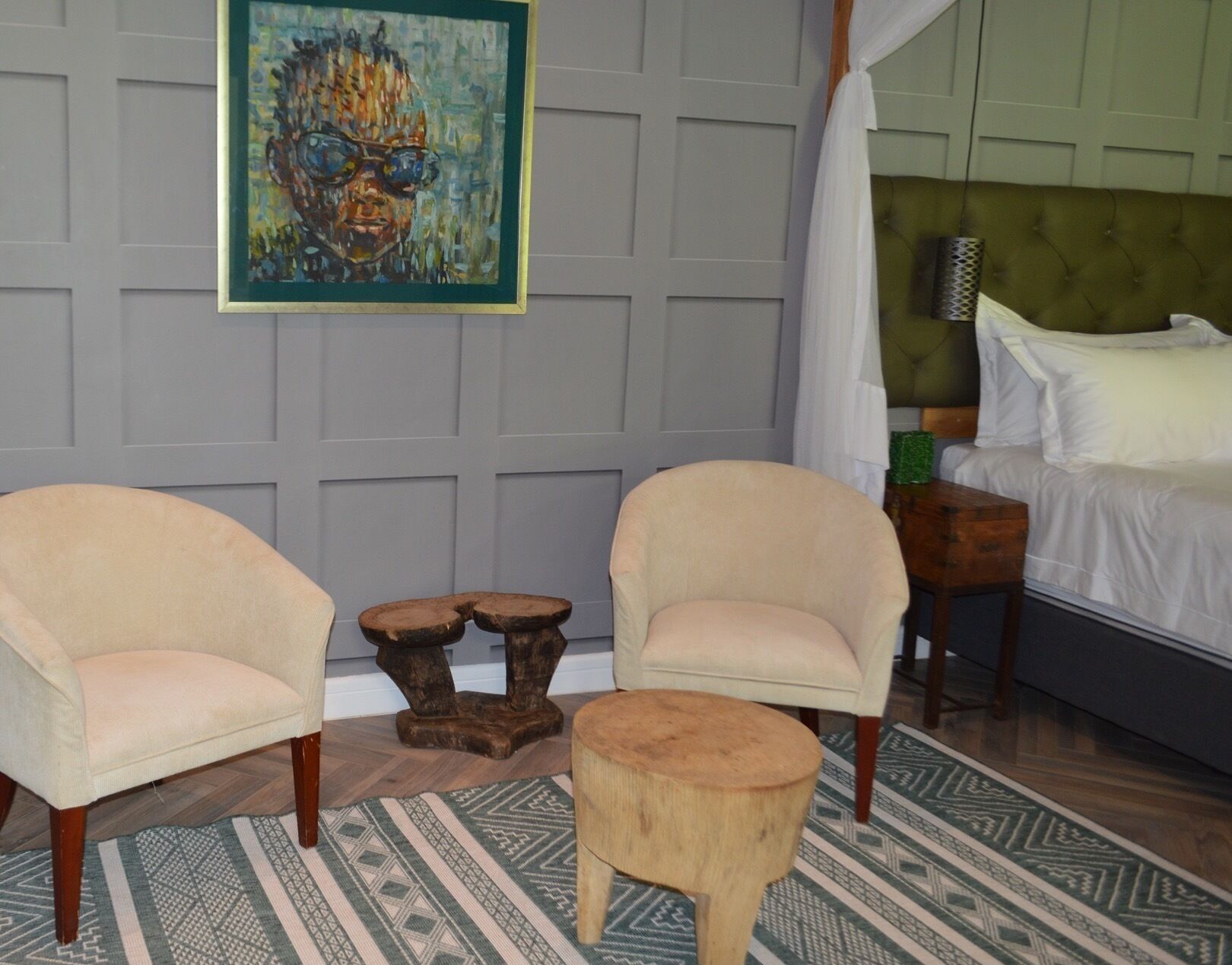 Een slaapkamer met twee fauteuils, een houten kruk, een bed met witte lakens, een groen hoofdeinde en een schilderij aan de muur.