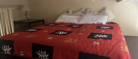 A bed with a red and black patterned bedspread, white pillows, and a small bedside table with a lamp.