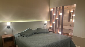 Una cama bien hecha con una colcha verde acolchada, dos lámparas de noche, una mesita de noche de madera y un tocador con espejo e iluminación.