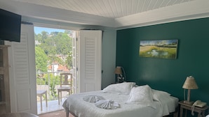 Un dormitorio con cama, una silla, una mesa, un cuadro y vistas al exterior.