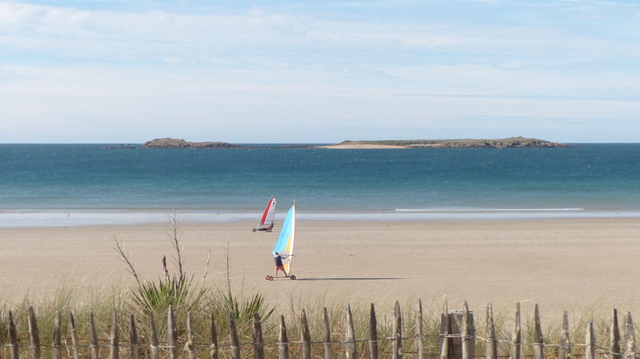 A sandy beach with a wooden fence in the foreground and a clear view of the sea and distant islands.