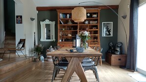 Un coin repas avec une table en bois, des chaises en métal, un luminaire en fibres tressées et une bibliothèque remplie de livres et d’objets décoratifs.