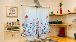 Ett kök med en dubbel diskho, bänkskivor i trä, en hängande gardin med blommotiv och hyllor med köksredskap.