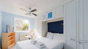 A hotel room with a bed, a chair, a desk with a computer, a window with a view of trees, and a ceiling fan. - Spacious 3BDR Kennington Home with Private Garden (London)