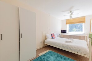 Ein Schlafzimmer mit Bett, Schreibtisch mit Computer, Fenster mit Jalousien und Deckenventilator.