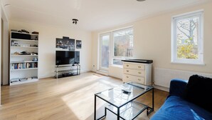 A modern living room with a flat-screen TV, a glass coffee table, a blue sofa, and a piano. - Spacious 3BDR Kennington Home with Private Garden (London)