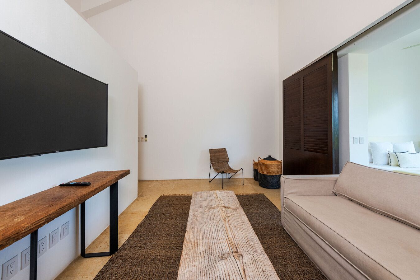 Una sala de estar moderna con un televisor de pantalla plana, un banco de madera, una silla marrón, un sofá beige y un ventanal con persianas.