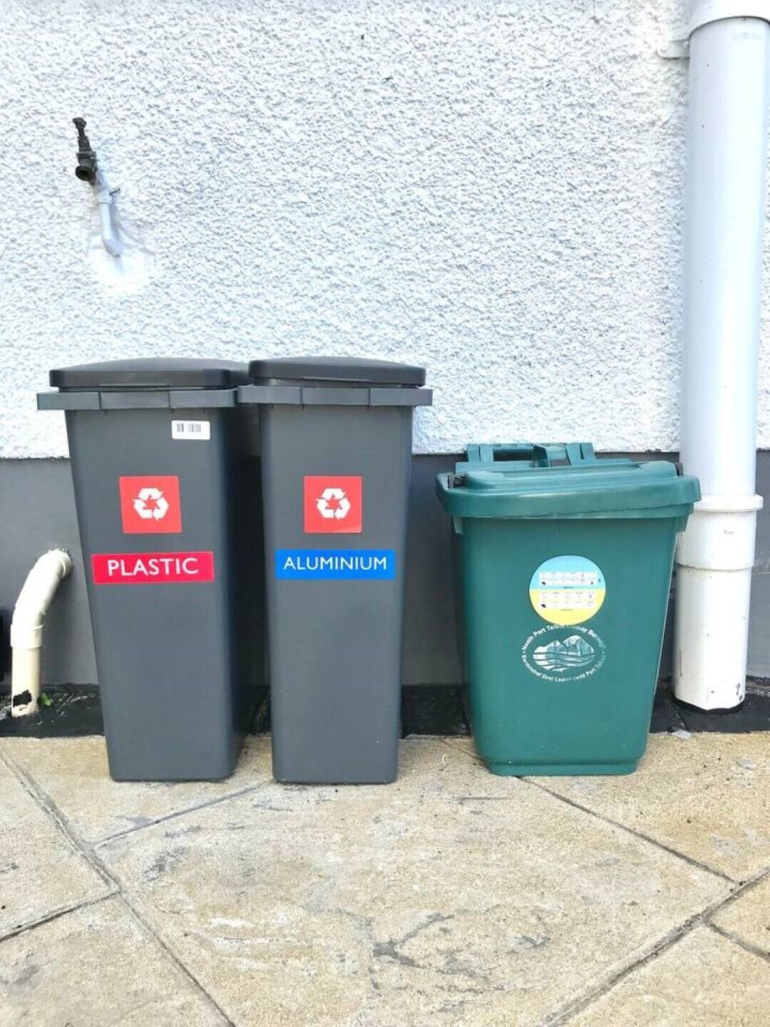 Tres dipòsits de reciclatge identificats per plàstic, alumini i residus general, col·locats davant d'una paret amb una canonada blanca.