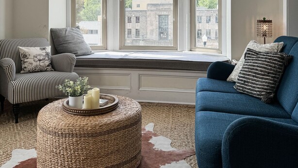 Een gezellige kamer met een erker, een gestreepte stoel, een blauwe bank en een geweven poef.