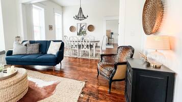Een woonkamer met een blauwe bank, een fauteuil met patroon, een ronde salontafel en een vloerkleed. In de achtergrond is een eethoek met een witte tafel en stoelen te zien.