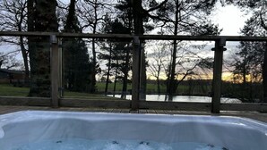 A hot tub with a wooden deck and metal railing, surrounded by trees and a view of a body of water. - Relaxing Woodland Retreat (Congleton)