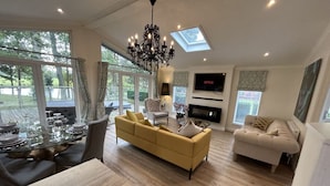 A spacious living room with a yellow sofa, a white sofa, a glass dining table set for six, and a fireplace. - Relaxing Woodland Retreat (Congleton)