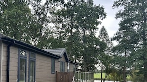 A cabin with a deck, surrounded by trees and a lake. - Relaxing Woodland Retreat (Congleton)