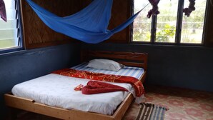 Una cama individual con estructura de madera, colchón blanco y manta roja. Hay una ventana con cortina azul y una alfombra estampada en el suelo.