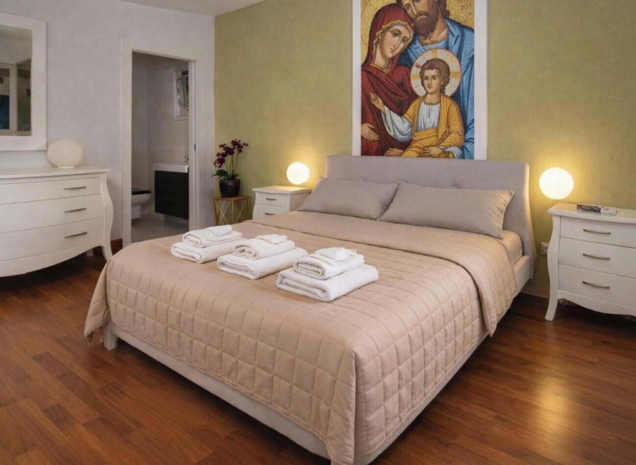 A bedroom with a large bed, two bedside tables, a nightstand, a painting on the wall, and a door leading to another room.