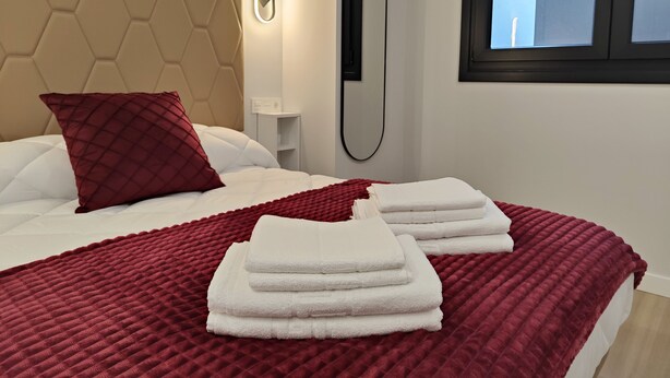 Ein ordentlich bezogenes Bett mit einer roten Steude, weißen Kissen und einer roten Tagesdecke. Auf dem Bett liegen gefaltete weiße Handtücher.