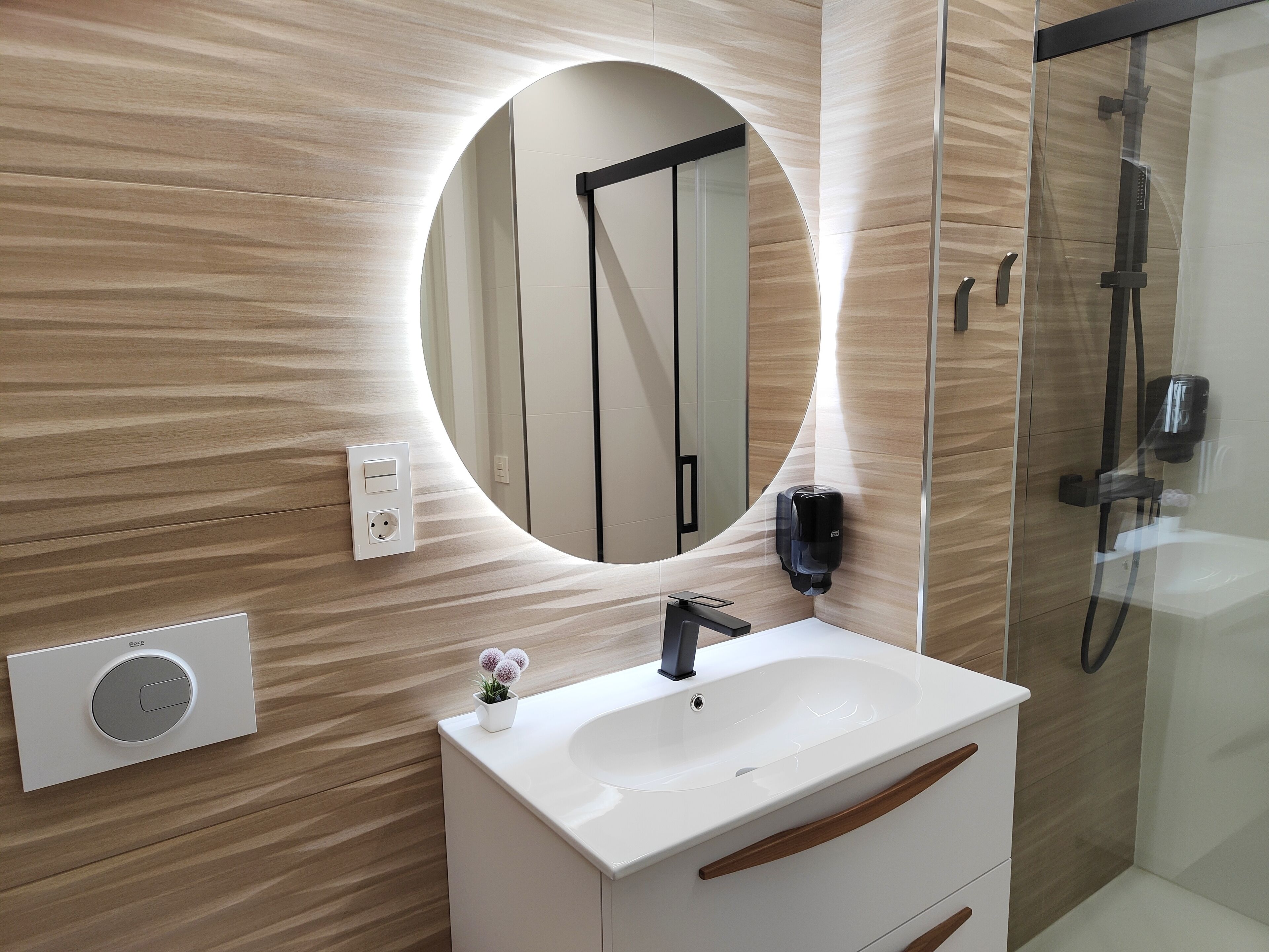 Ein modernes Badezimmer mit rundem Spiegel, Waschbecken in Weiß, Duschbrausehandstück und Handtuchspender.