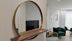 Ein modernes Wohnzimmer mit einem großen, runden Spiegel an der Wand, einem Holzregal darunter, einem Flachbildfernseher, einem grünen Sofa und einem hellen Boden.