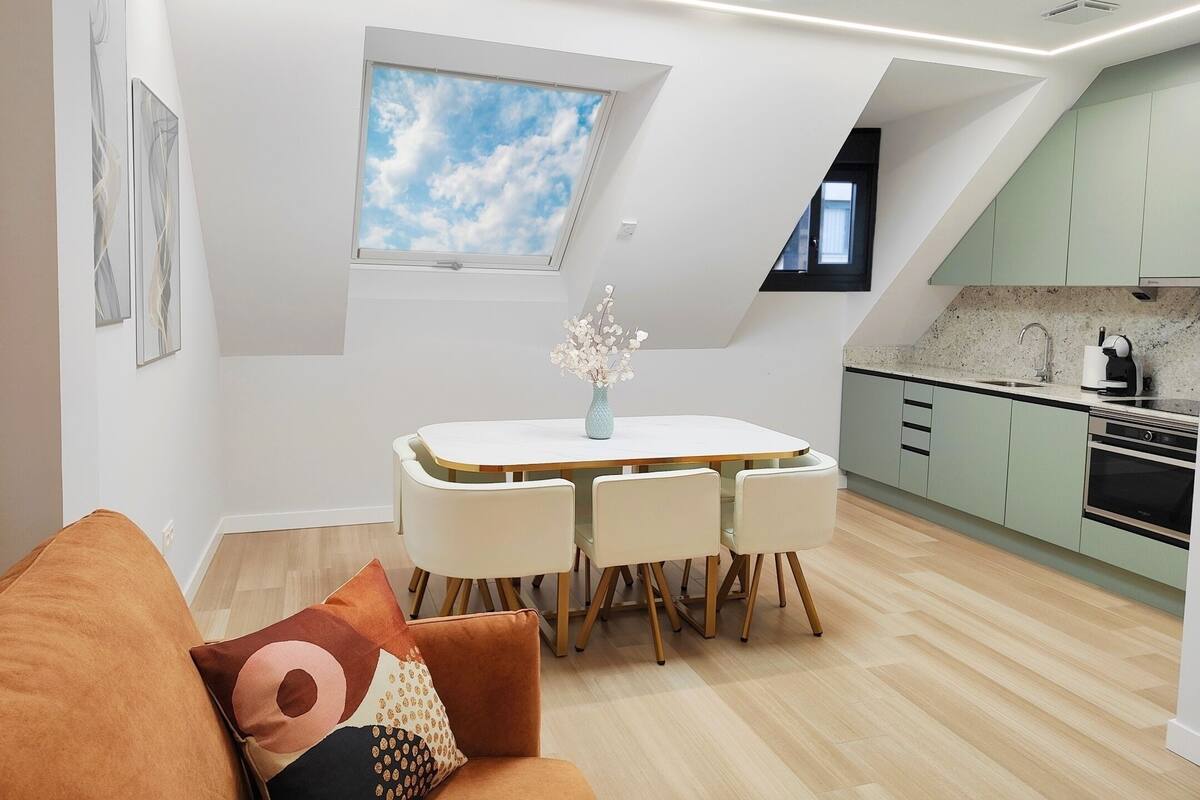 Une cuisine moderne dotée d’une lucarne, un coin repas avec une table et des chaises, et un salon avec un canapé et des coussins.