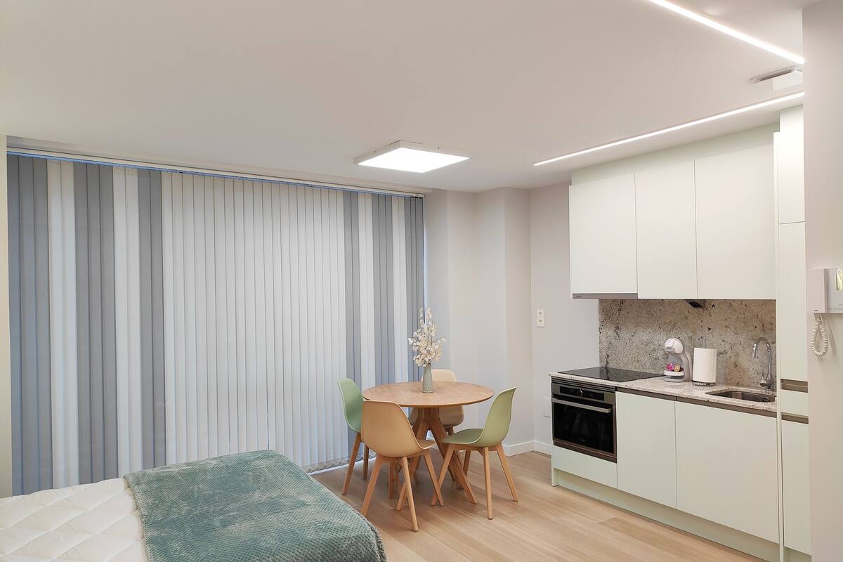 Une chambre d’hôtel moderne comprenant un lit, une table à manger avec des chaises, un coin cuisine avec une cuisinière et des placards, ainsi qu’une fenêtre équipée de stores verticaux.