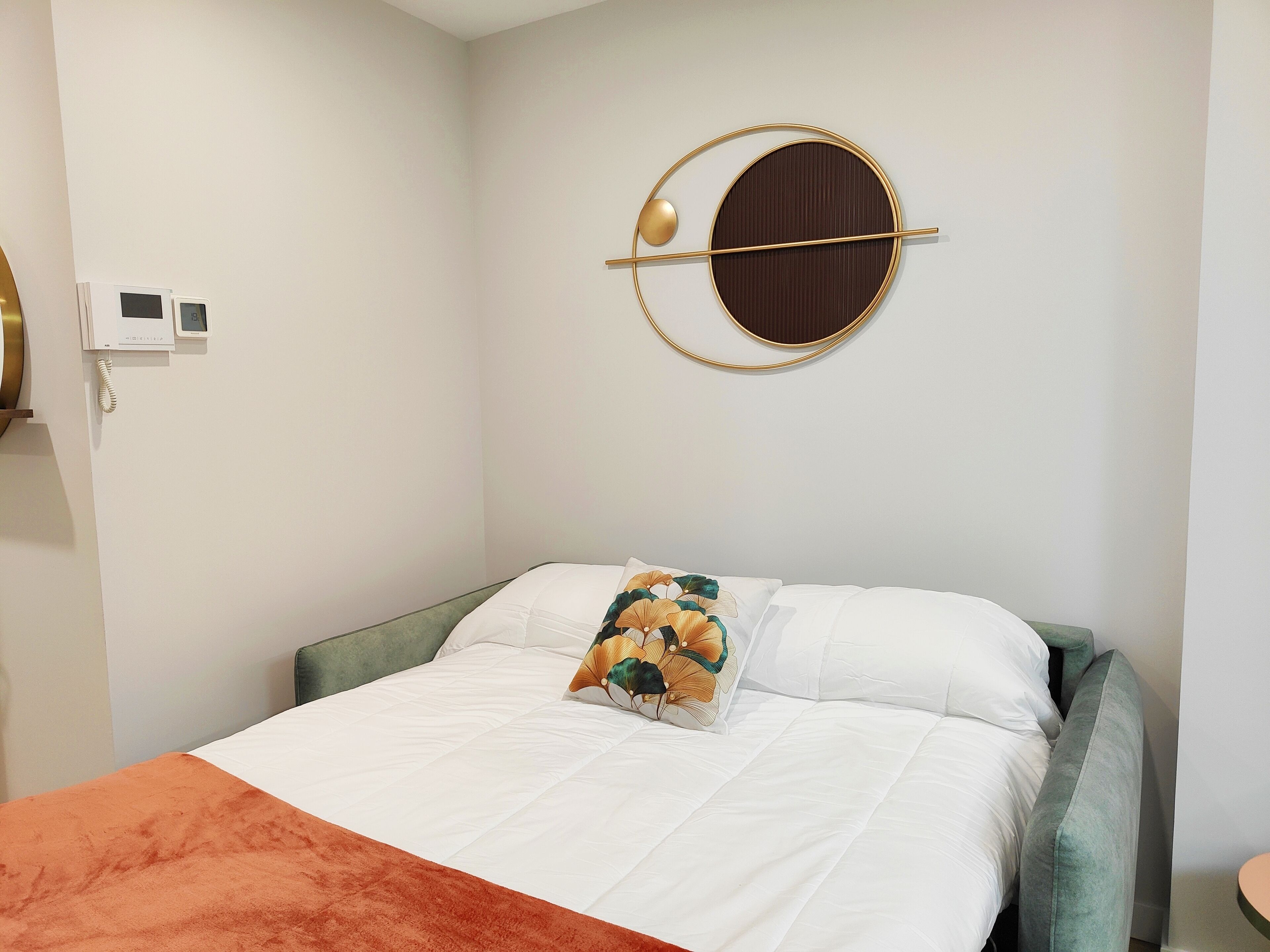 A neatly made bed with white bedding and a decorative pillow, a round wall decoration, and a telephone on the wall.
