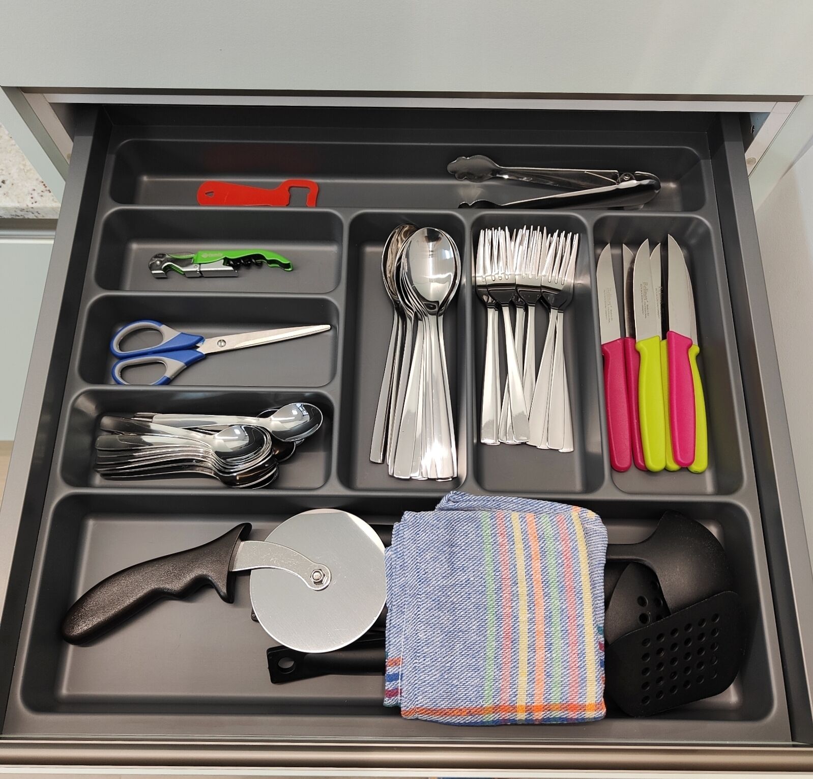 A drawer with compartments containing various kitchen utensils, including scissors, a knife, a spoon, and a pair of tongs.