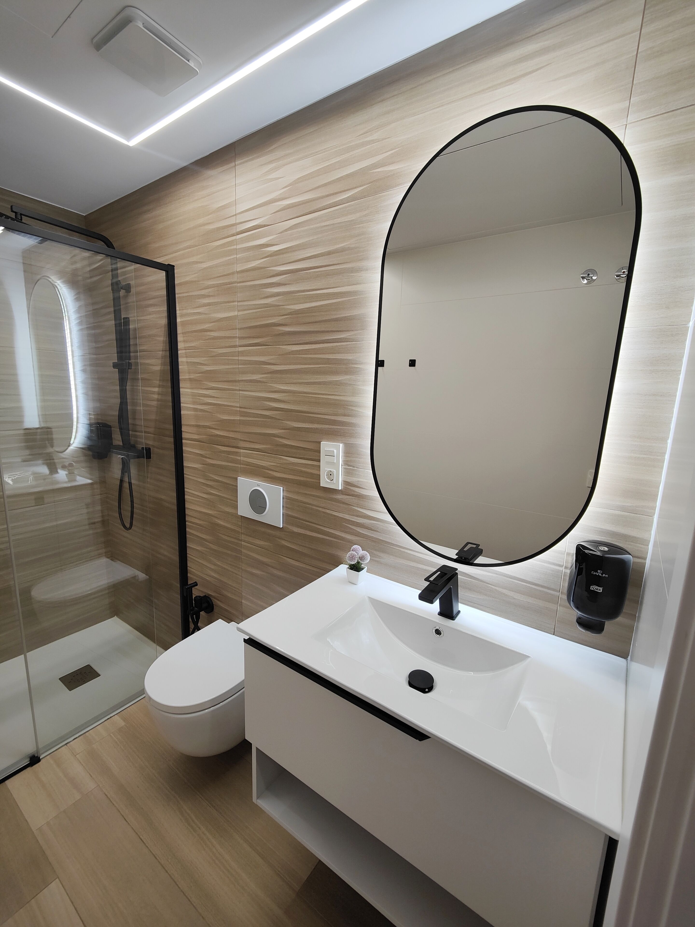 Un baño moderno con ducha envuelta en vidrio, un lavamanos blanco con grifo negro y un espejo ovalado grande.