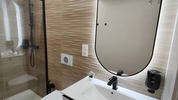 Un baño moderno con ducha envuelta en vidrio, un lavamanos blanco con grifo negro y un espejo ovalado grande.