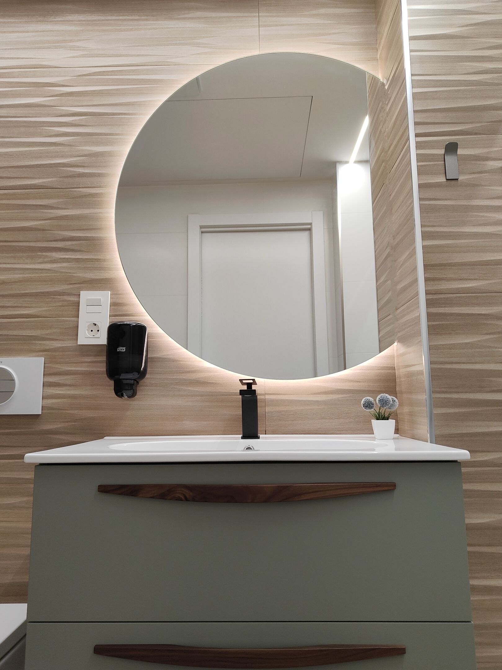 A bathroom with a round mirror, a sink, a faucet, a soap dispenser, a potted plant, and a door.