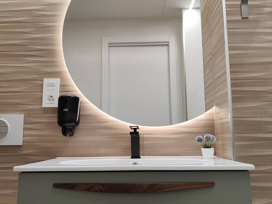 A bathroom with a round mirror, a sink, a faucet, a soap dispenser, a potted plant, and a door.