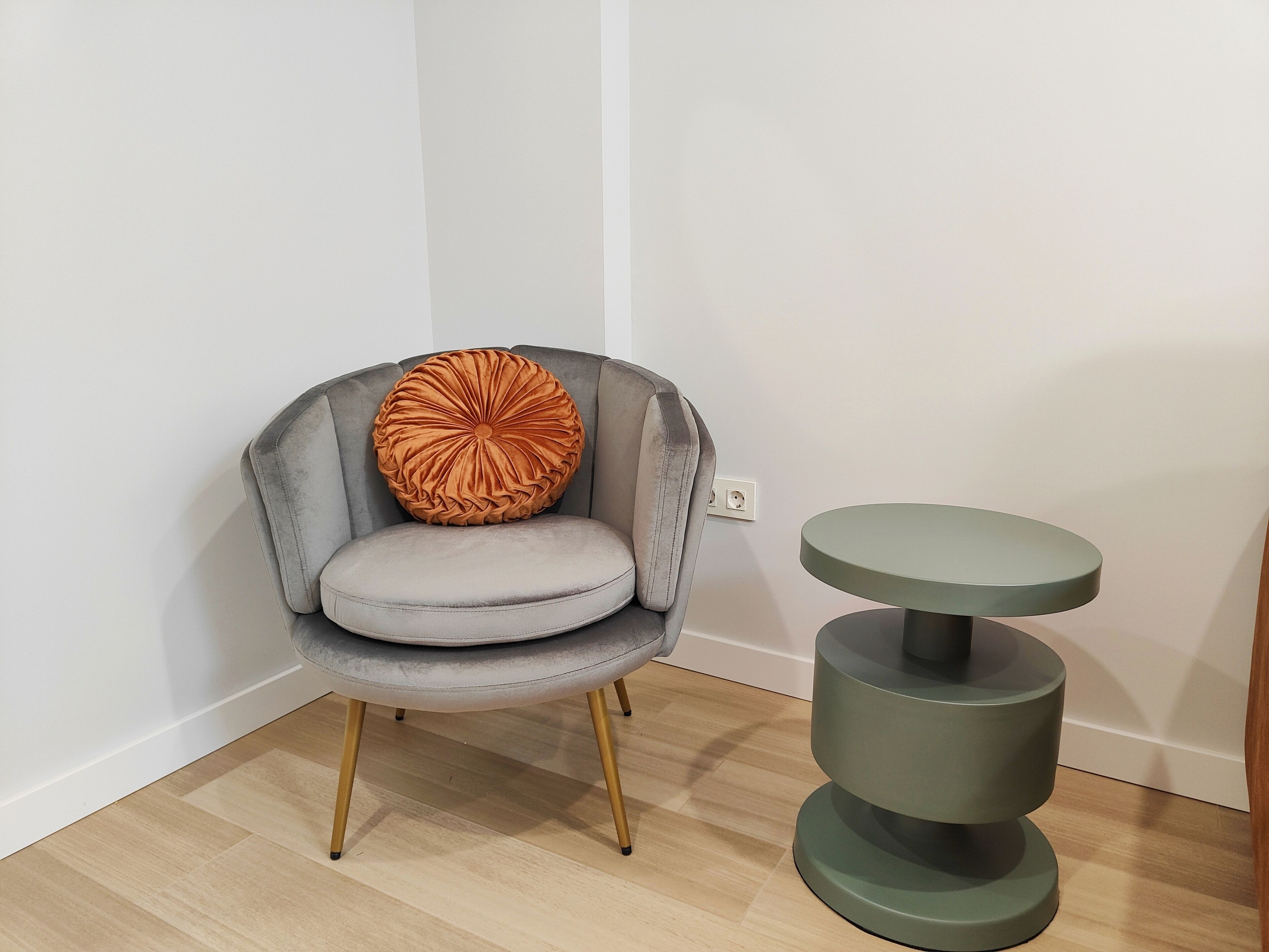 A grey upholstered armchair with a round, textured orange pillow, positioned next to a green, multi-tiered side table.