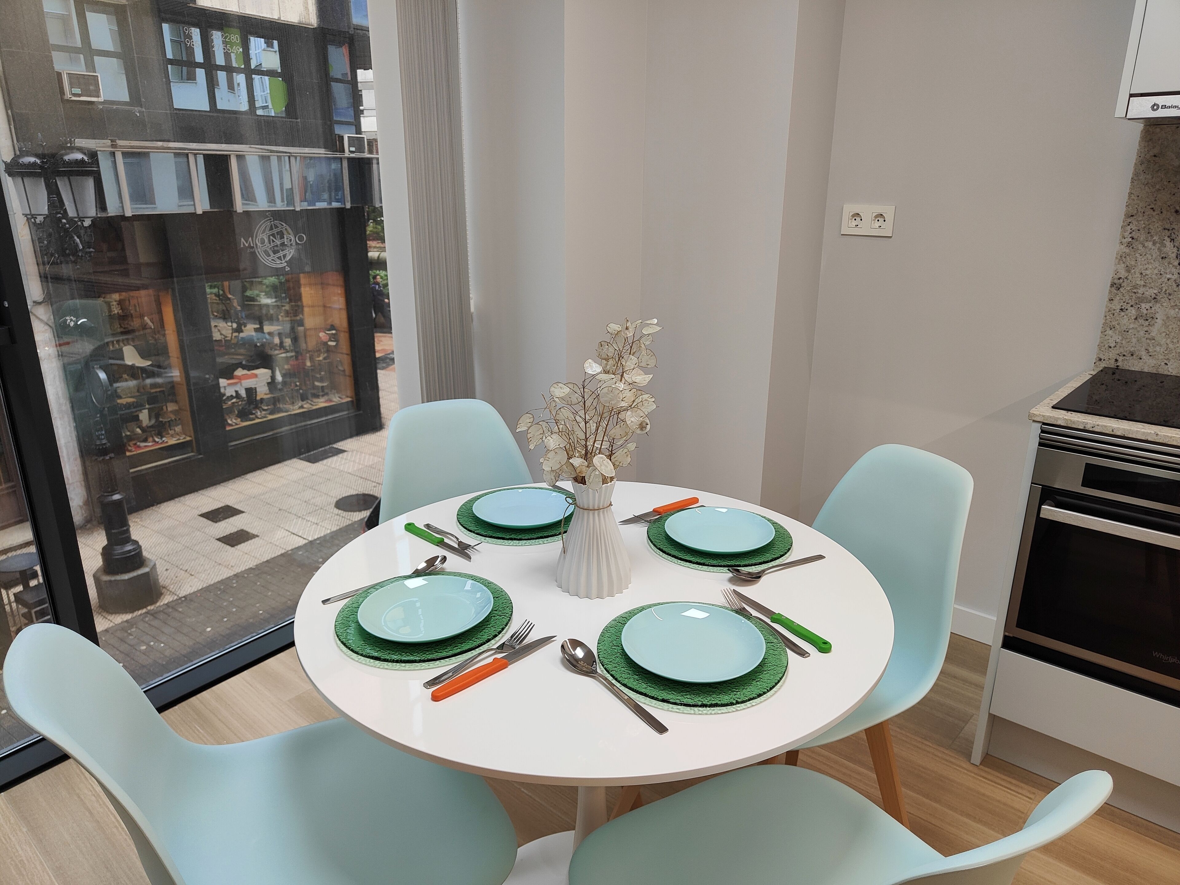 Un comedor con una mesa redonda dispuesta para cuatro personas, sillas azul claro y un jarrón con flores secas.
