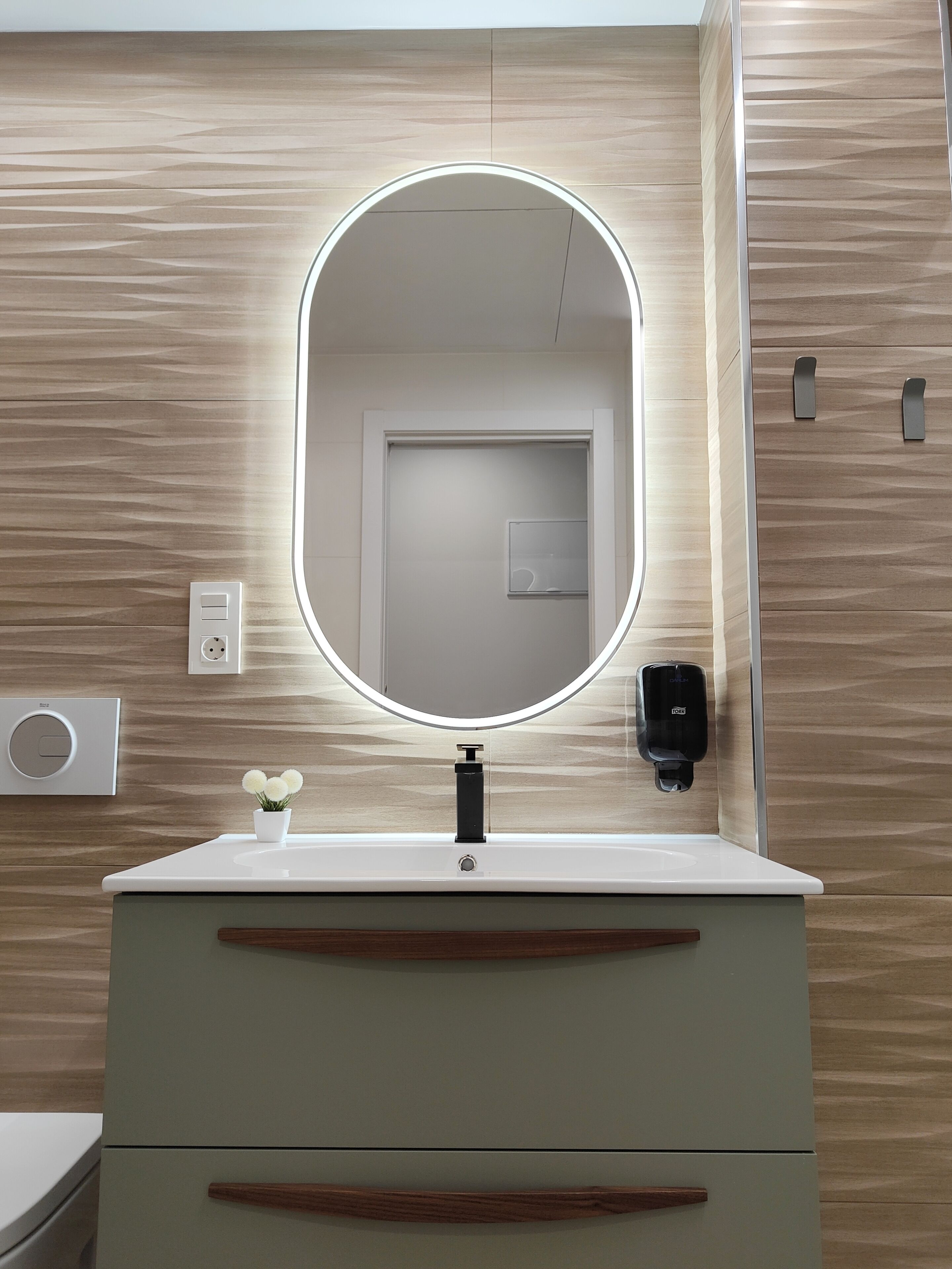 Une salle de bain avec un meuble-lavabo blanc, un miroir rond doté d’un éclairage intégré, une petite plante en pot et un distributeur de savon.