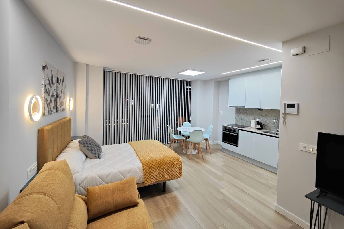 Une chambre d’hôtel moderne comprenant un lit, un canapé, un coin repas avec une table et des chaises, une cuisine équipée de placards et d’appareils électroménagers, et un téléviseur posé sur un meuble.