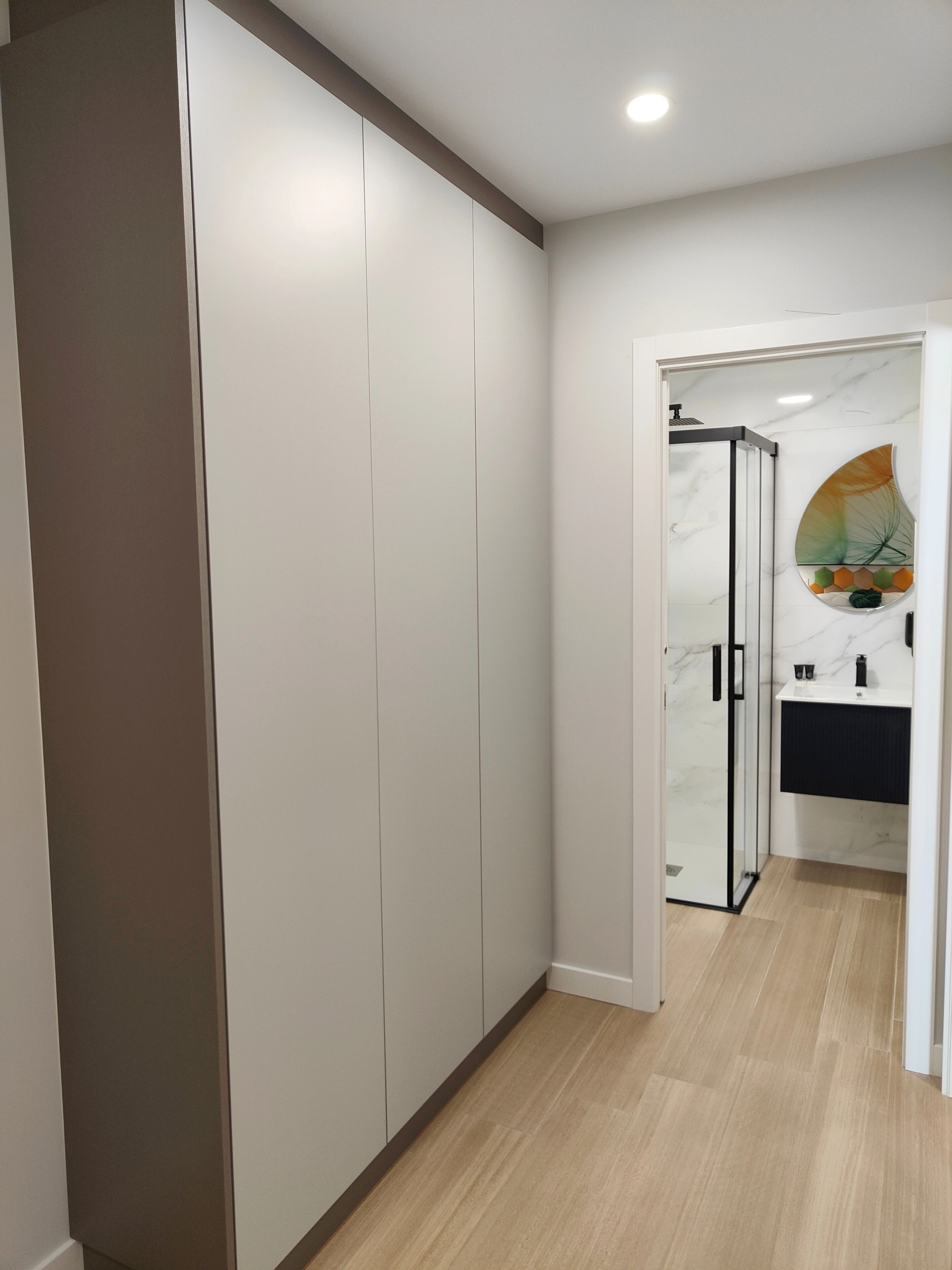 Un pasillo con un armario, cabina de ducha con mampara de vidrio y un baño con lavamanos y espejo.