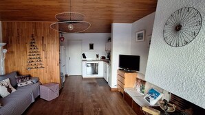 Een gezellige woonkamer met een bank, een klein tafeltje en een televisie. Achtergrond is een keuken met een koelkast en een magnetron.