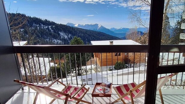 Een balkon met twee gestreepte stoelen en een bijzettafeltje met een dienblad, met uitzicht op een besneeuwd berglandschap.