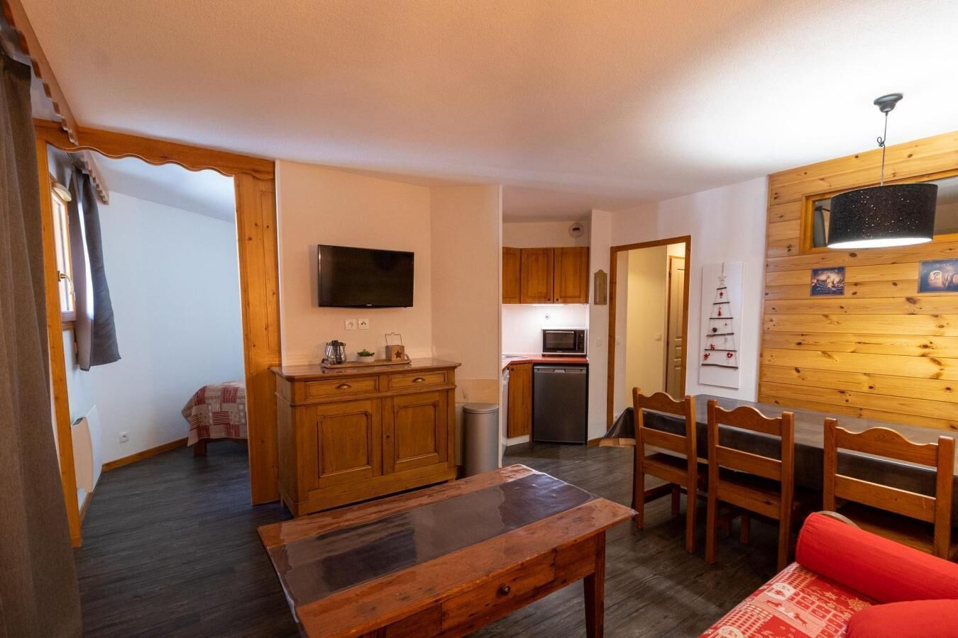 A compact living space with a wooden dining table, a red sofa, a kitchen area with wooden cabinets, a flat-screen TV, and a small dining table.