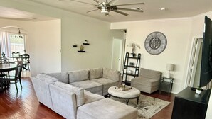 A living room with a sectional sofa, a round ottoman, a coffee table, a TV stand, and a ceiling fan.