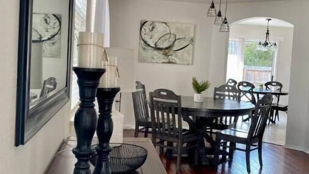 A dining area with a wooden table, chairs, and a chandelier. There are abstract paintings on the walls and a window providing natural light.