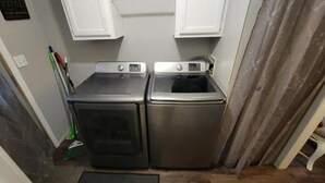 A compact laundry area with two stainless steel washing machines and a dryer, located in a small room with white cabinets above.