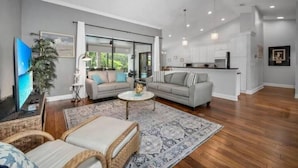 A spacious living room with a large sofa, a coffee table, and a TV. There is a kitchen area with white cabinets and a dining table.