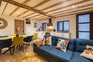 Ein modernes Wohnzimmer mit einem blauen Sofa, Holzbalkendecke, einem Essbereich mit Holztisch und -stühlen sowie einer Küche im Hintergrund.