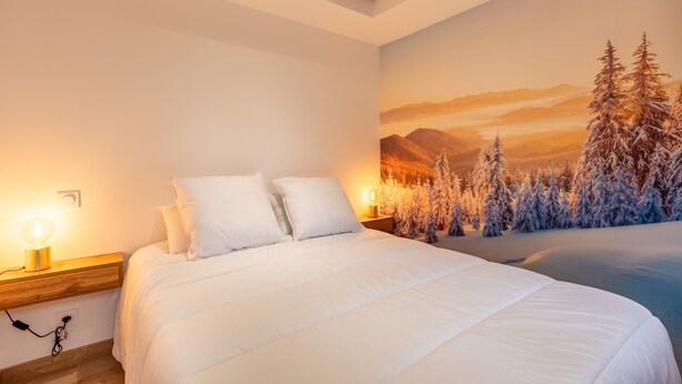 Un dormitorio con cama, ropa de cama blanca, una mesita de noche con lámpara y un mural en la pared con paisaje de un bosque nevado.