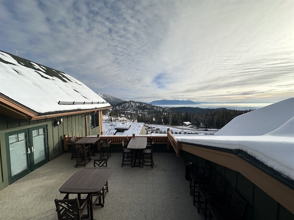 A covered outdoor patio with tables and chairs, a view of snow-covered mountains, and a building with green siding.