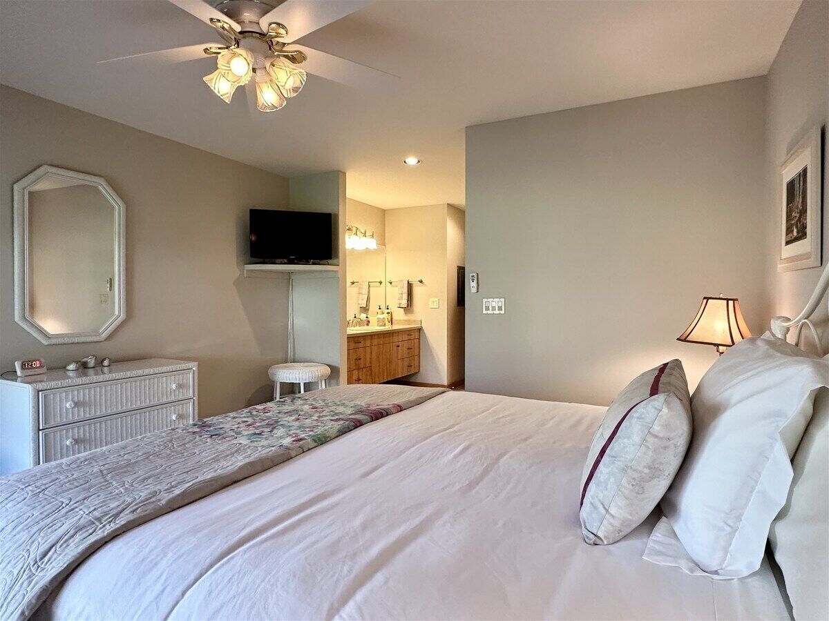 A bedroom with a bed, a dresser, a vanity, a ceiling fan, and a television.