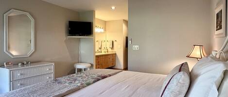 A bedroom with a bed, a dresser, a vanity, a ceiling fan, and a television.