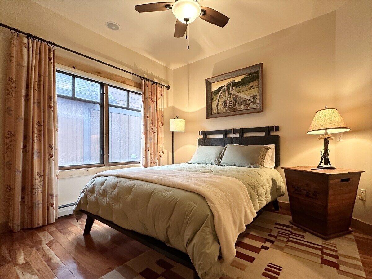 Une chambre à coucher avec un lit, un ventilateur de plafond, une table de chevet avec une lampe, une fenêtre avec des rideaux et un tableau accroché au mur.