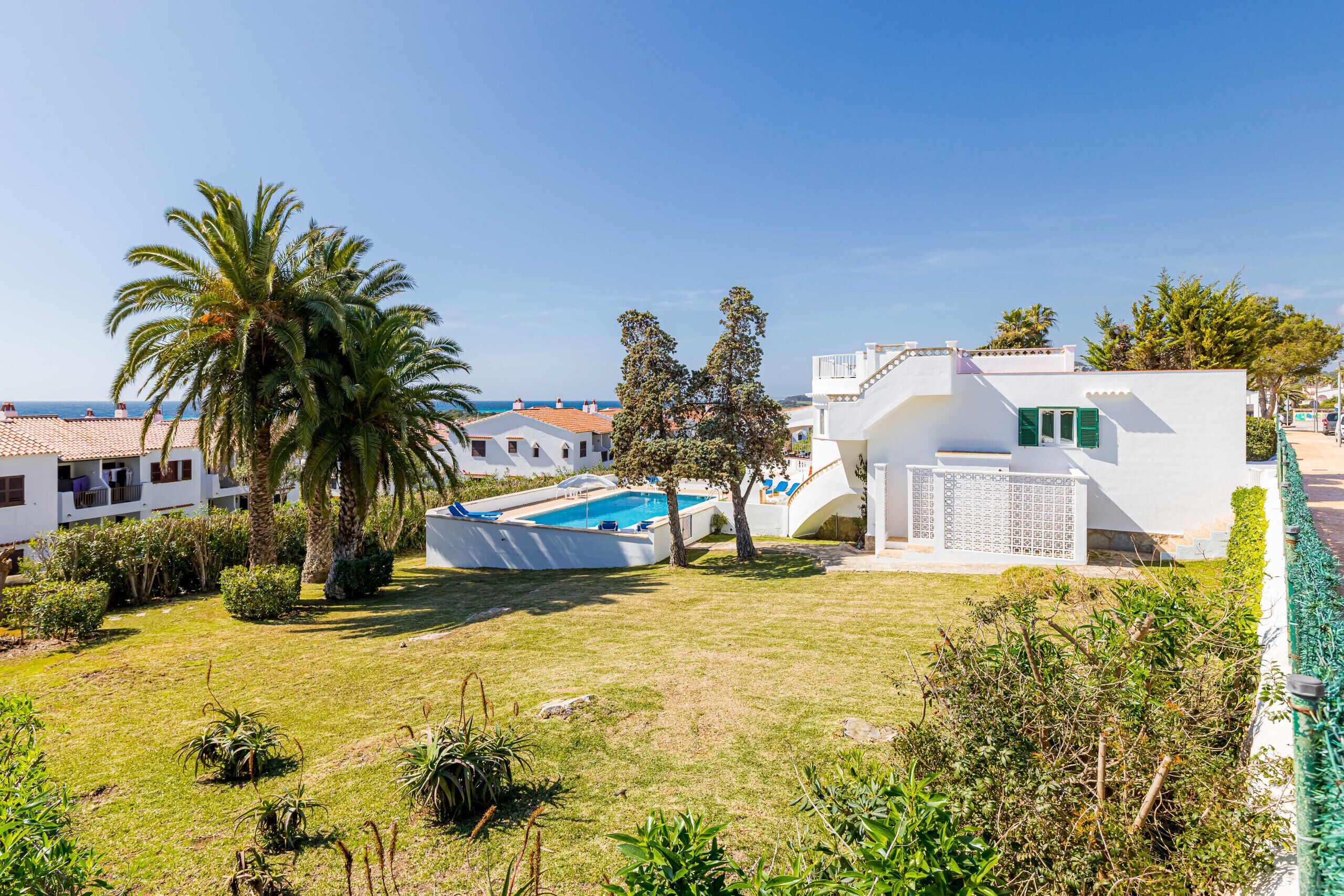 A white villa with a swimming pool, surrounded by greenery and a clear view of the sea.