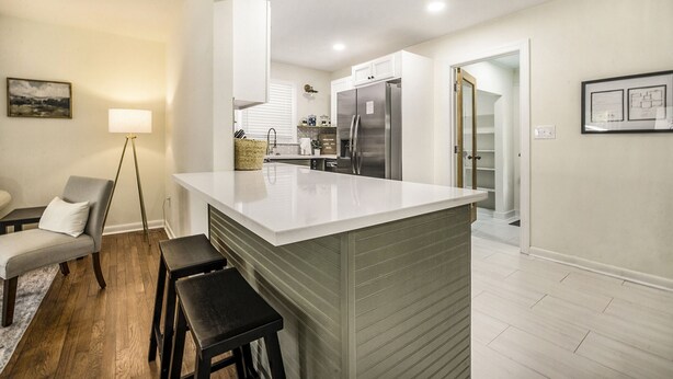 A modern kitchen with a central island, white cabinetry, and stainless steel appliances. There is a dining area with a chair and a lamp.